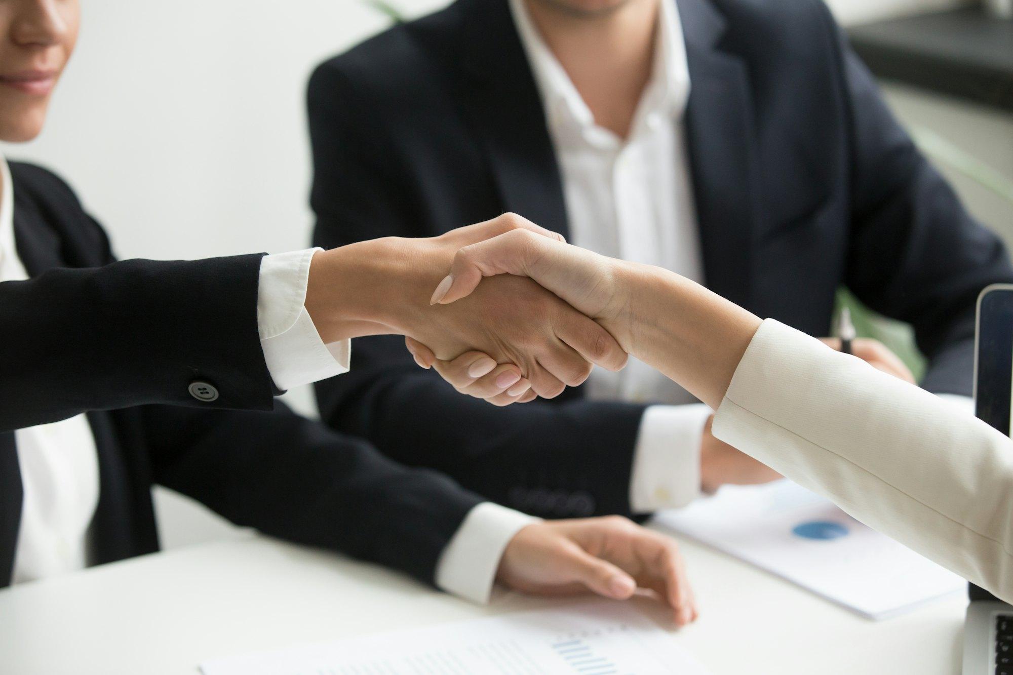 Two business partners in suits shaking hands over a desk